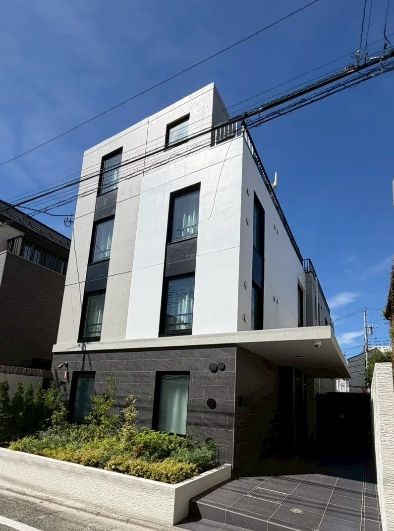 現代的な白と黒の三階建ての住宅外観。建物前に緑の植物と花壇。晴れた青空。