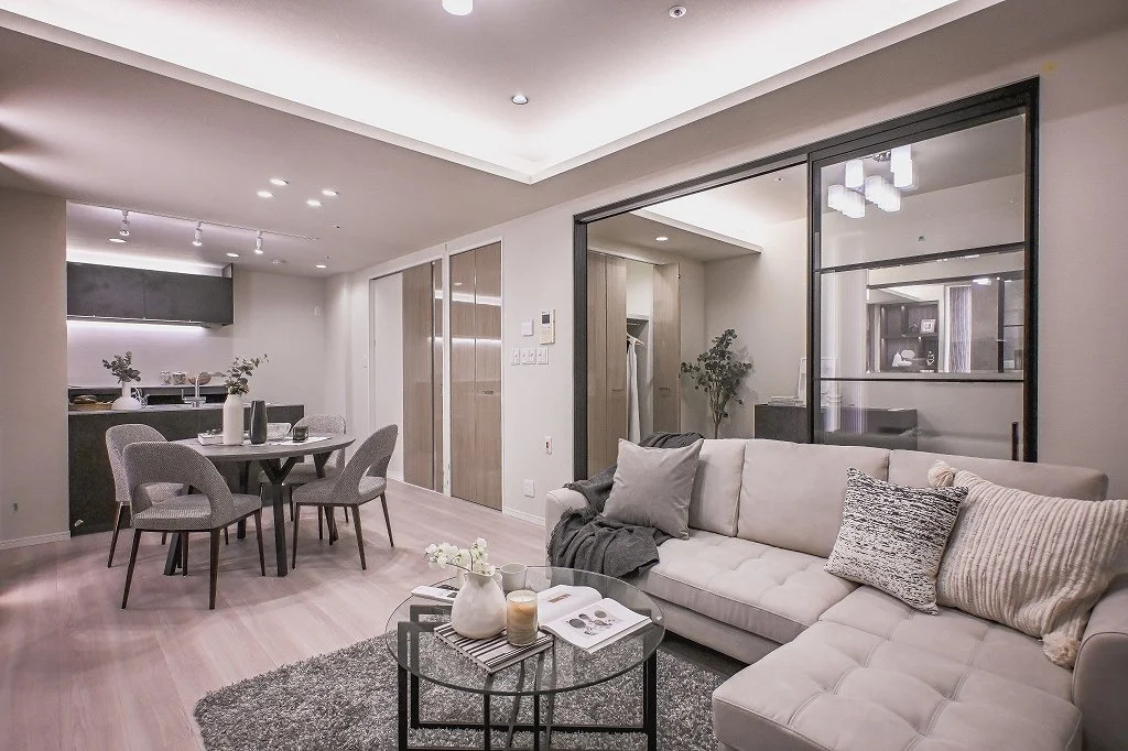 モダンなリビングルームとダイニングエリアの画像。白いソファ、ガラスのコーヒーテーブル、ダイニングテーブルと椅子、キッチンエリア、壁に窓やドア、柔らかい照明が見える。