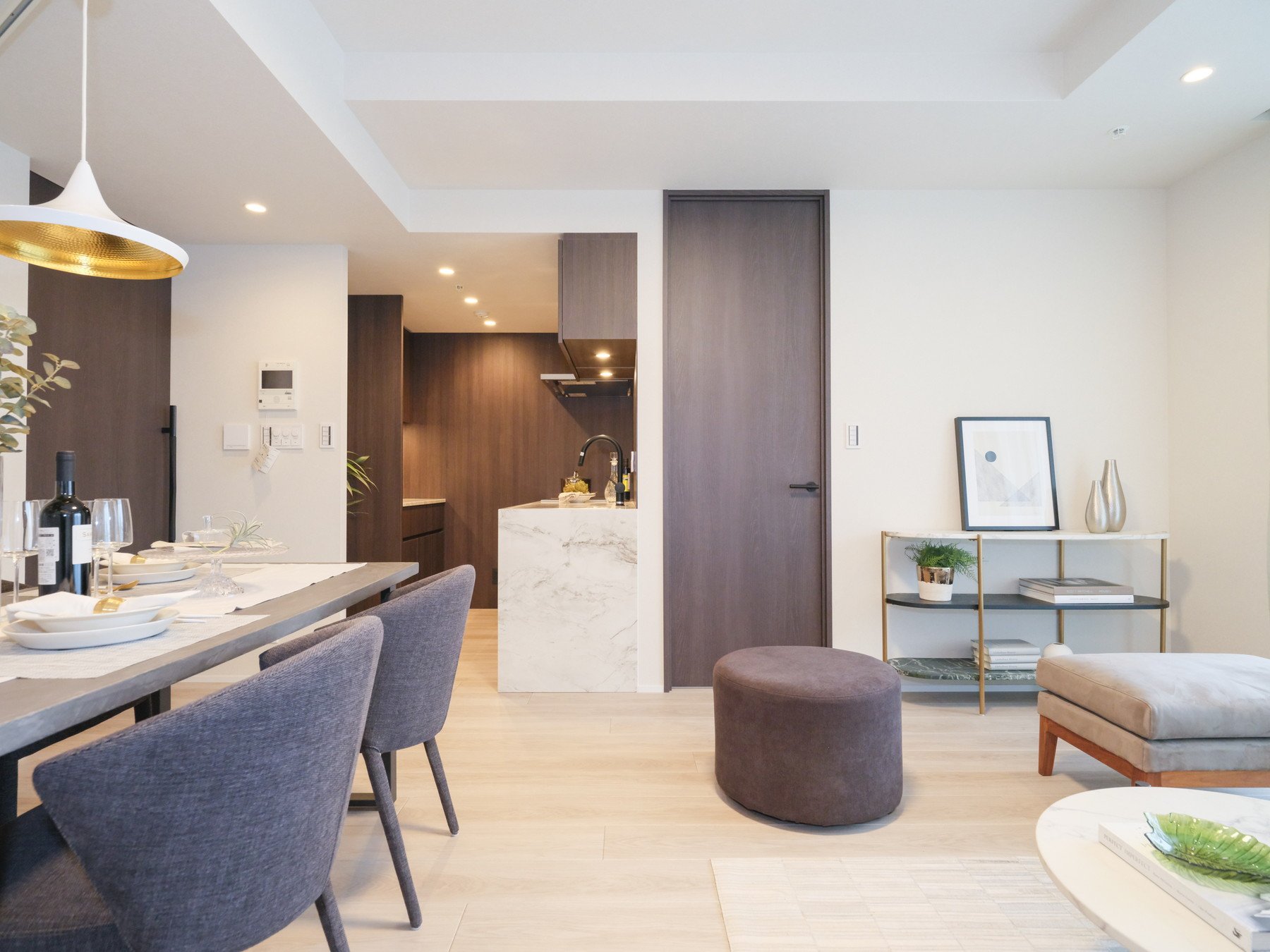 モダンなリビングルームとダイニングエリアの画像。ダイニングにはテーブルと灰色の椅子、リビングには丸いクッションの座席と棚に装飾品や本が置かれている。キッチンは背景に見える。