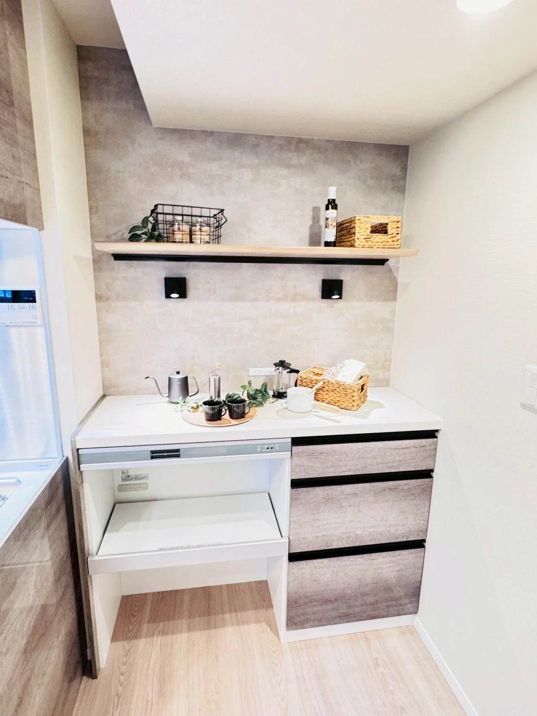 コンパクトなキッチンの一部、白いカウンターと黒いハンドルの引き出し棚、上にはコーヒー関連用品と植物が飾られている。