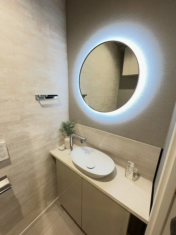 シンプルで現代的な洗面所に、白い円形の洗面ボウルと鏡、LEDライト付きの丸い鏡、少しの植物とハンドソープがあります。