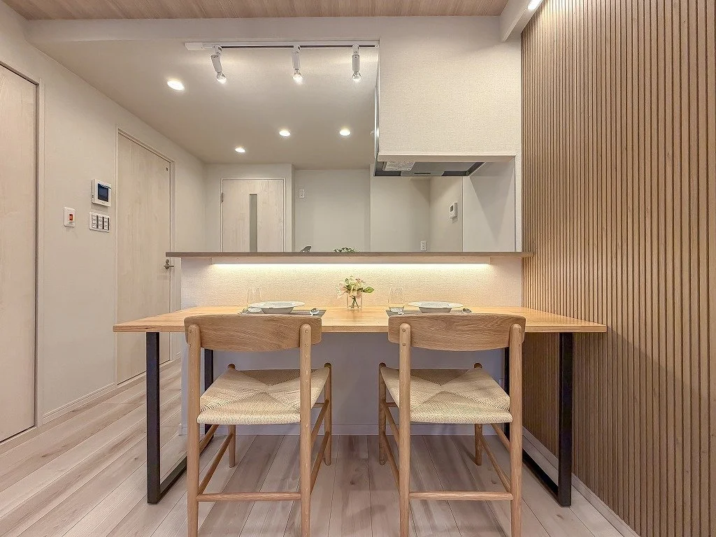 木製のテーブルと椅子、壁は白と木目調のデザイン。キッチンエリアのカウンター越しにダイニングが見える。