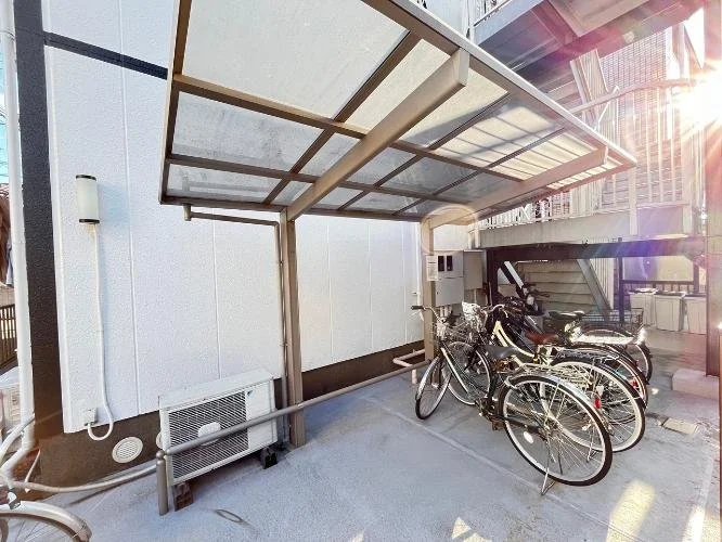 屋根付きのバイク・自転車駐輪場に数台の自転車が駐輪されている。背景に建物と階段が見える。