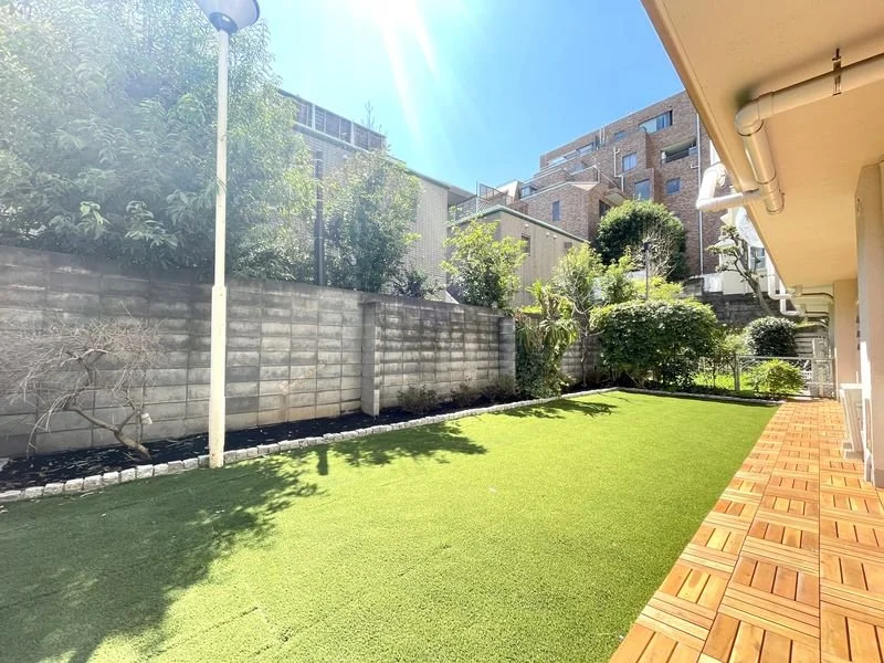 緑の芝生と花壇、木と高層マンションの背景が見えるバルコニーの庭。日差しが強い晴れた日。