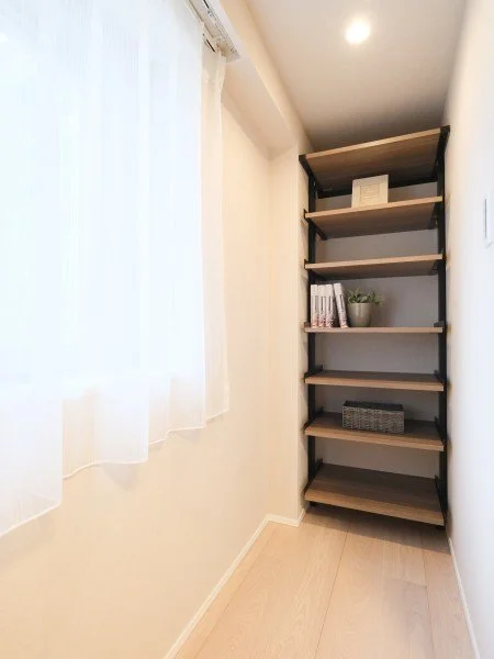 白い壁とカーテンのある部屋に、木製と金属の本棚が置いてあり、本棚には本や観葉植物、収納ボックスが備えられている。