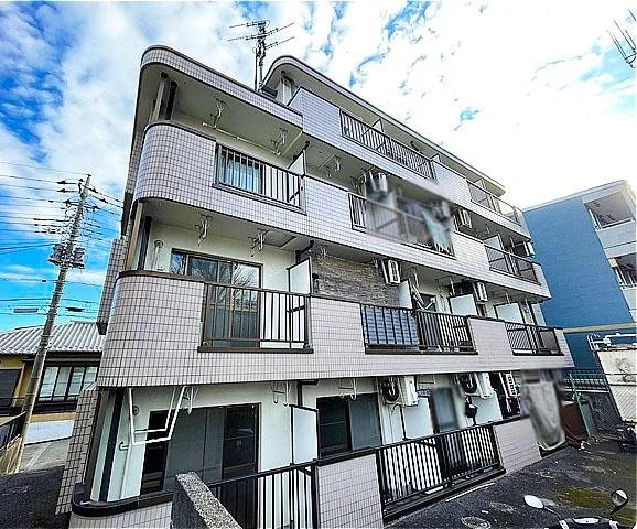 三階建ての白いタイル張りのアパートメント建物。各階にバルコニーと窓、エアコンが設置されている。空と一部の電線が見える。
