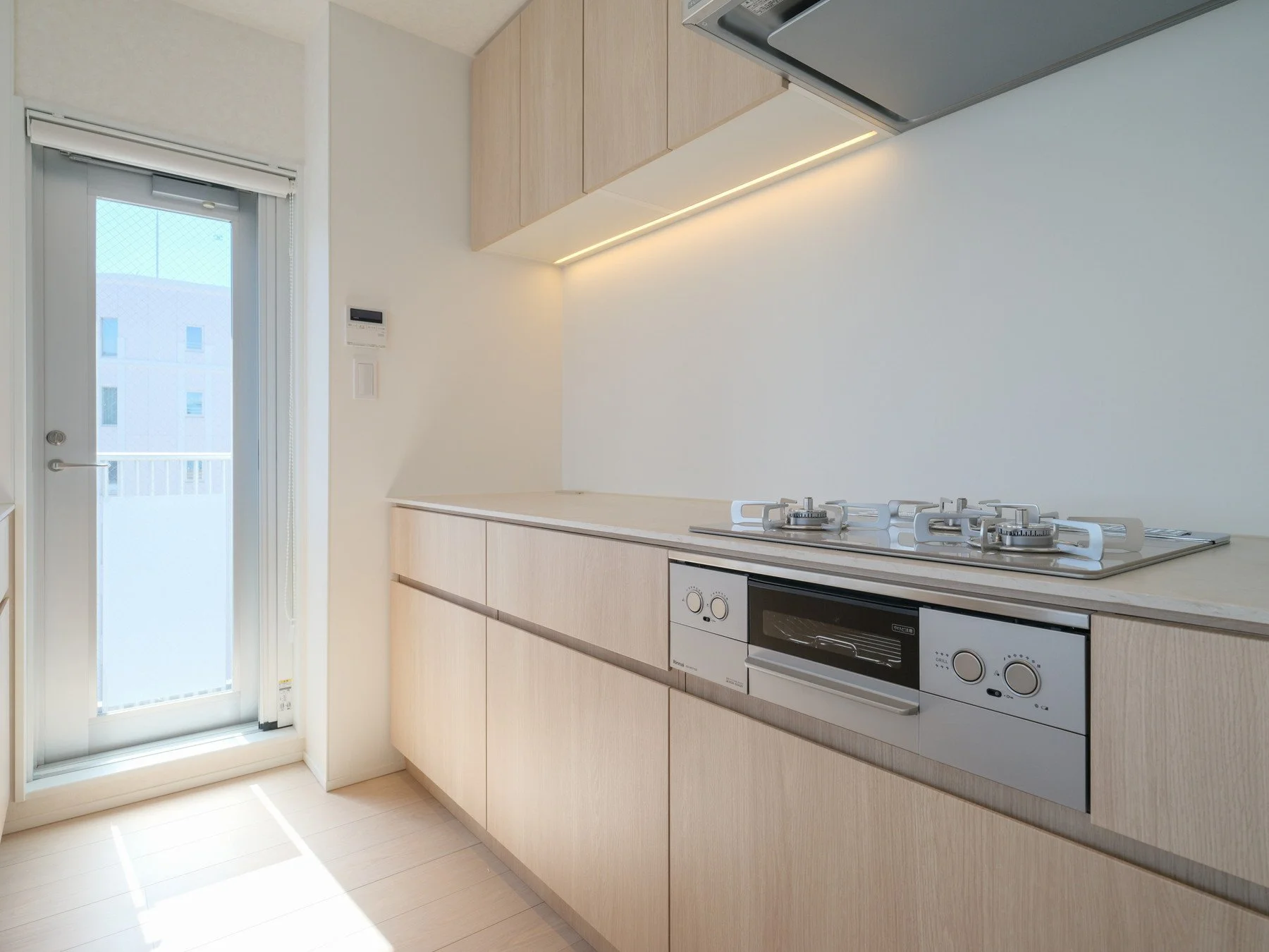 明るいキッチンの内部、白い壁と木材のキャビネット、ガスコンロとオーブン付きのカウンター、背面には窓とバルコニーのドア