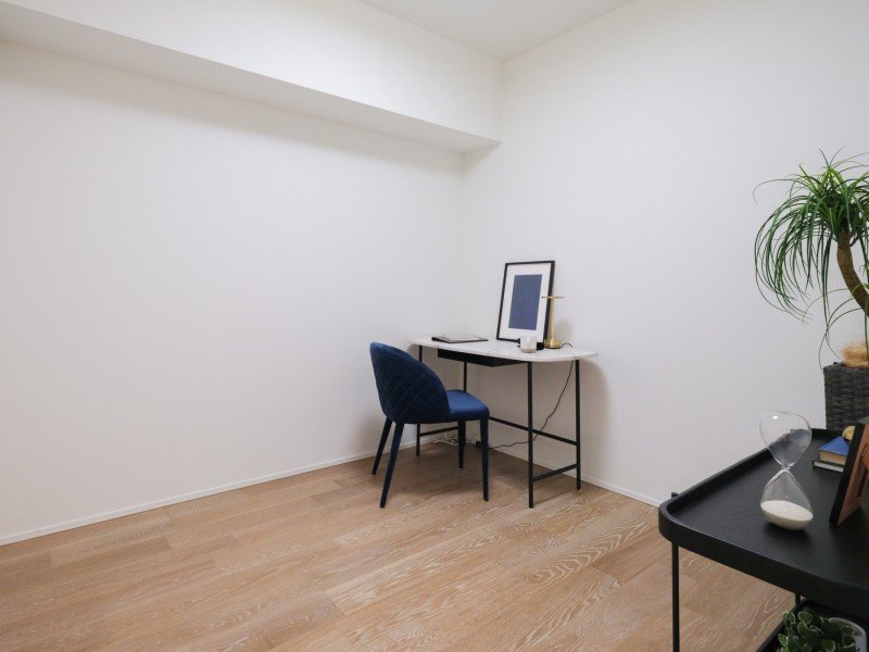 白い壁の部屋に茶色の床、机と青い椅子、パソコン、観葉植物、砂時計、いくつかの本や装飾品が置かれている