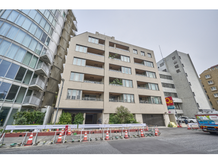 都市部の中層の建物と向かい側の道路と駐車場、工事用のバリケードや車両が見える風景