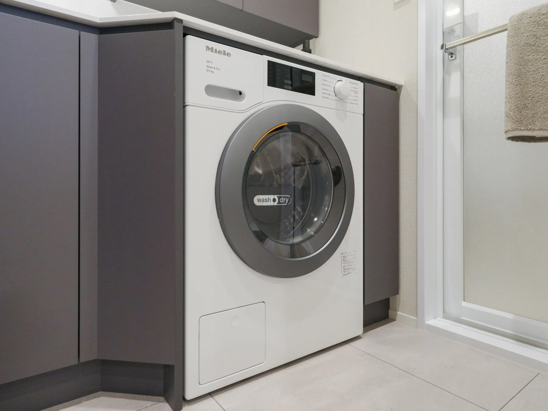 白い洗濯機が灰色のキャビネットの横に設置されている。洗濯機には「wash」と「dry」のラベルがあり、丸いドアから中の洗濯物が見える。