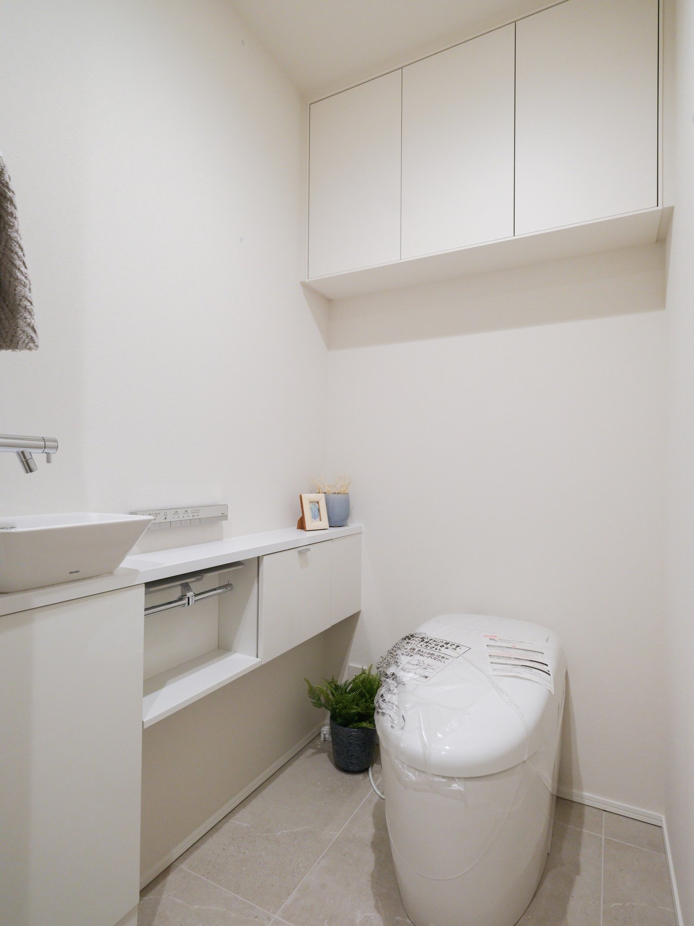 白い壁と収納棚があるトイレの空間。トイレは新しいもので、袋がかけられている。棚の上には小さな写真立てと植木鉢が置かれている。