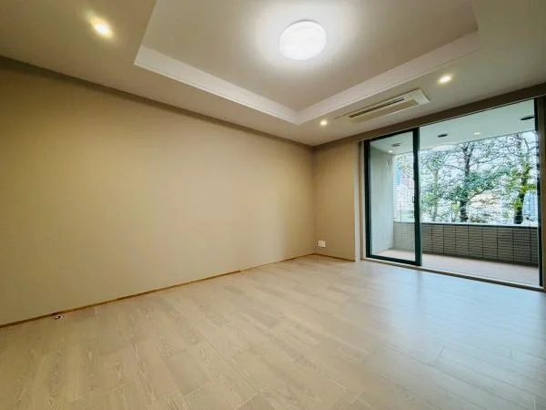 空の広い部屋で、木目調の床と白い壁、天井に照明があり、ガラスのスライドドアを通じてバルコニーと外の景色が見える。