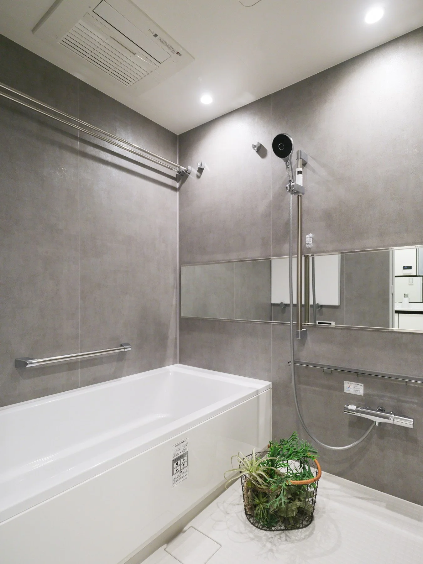 バスルームの壁にシャワーとバスタブ、鏡、植物の飾りがあり、天井には換気扇と照明がある。