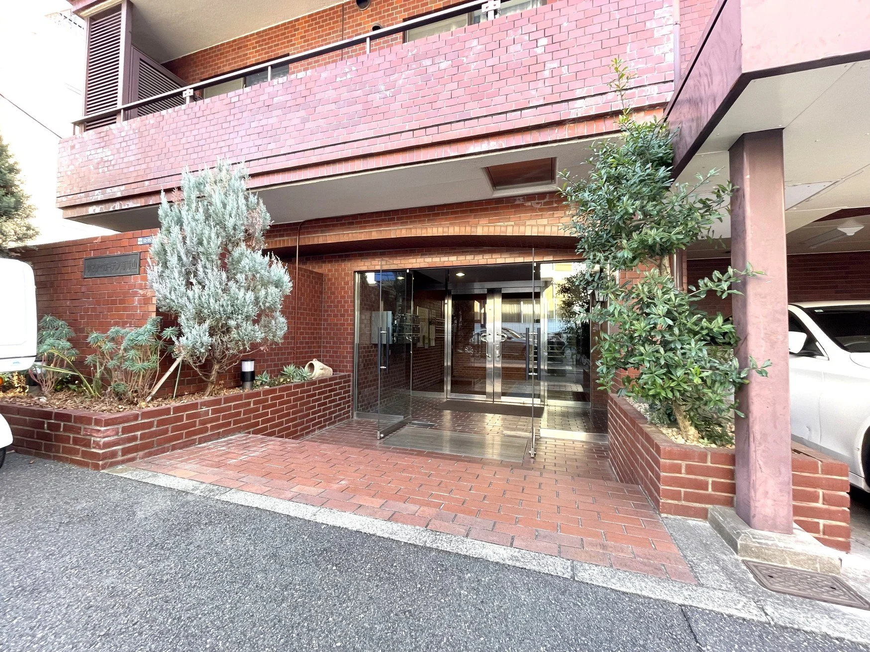 赤レンガの建物の入り口で、ガラスドアには金属の手すりがついています。前には植物の植え込みがあり、左右に木が植えられています。