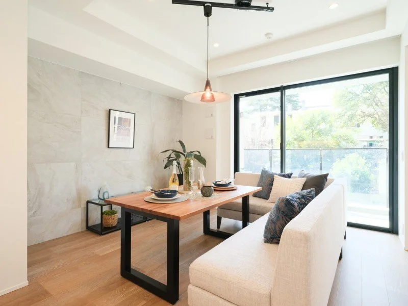 窓からの自然光が差し込むリビングダイニングルームの写真。テーブルの上に花と食器が置かれ、ソファと背もたれのクッションと飾りがあり、シンプルでモダンなインテリアです。