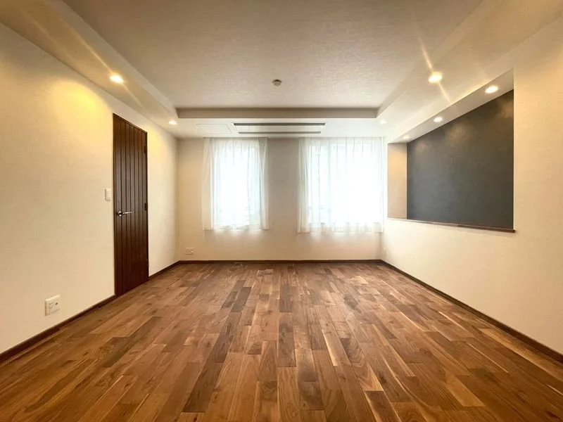 明るい空間のリビングルーム。木製の床と白い壁、窓に白いカーテンがあり、黒板のような黒い壁面もある。