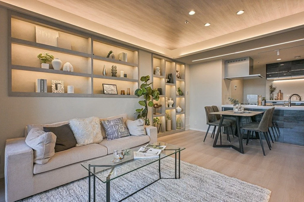 モダンなリビングルームとダイニングキッチンのインテリアで、ソファ、ガラスのコーヒーテーブル、収納棚に飾り物や本、植物が配置されている。