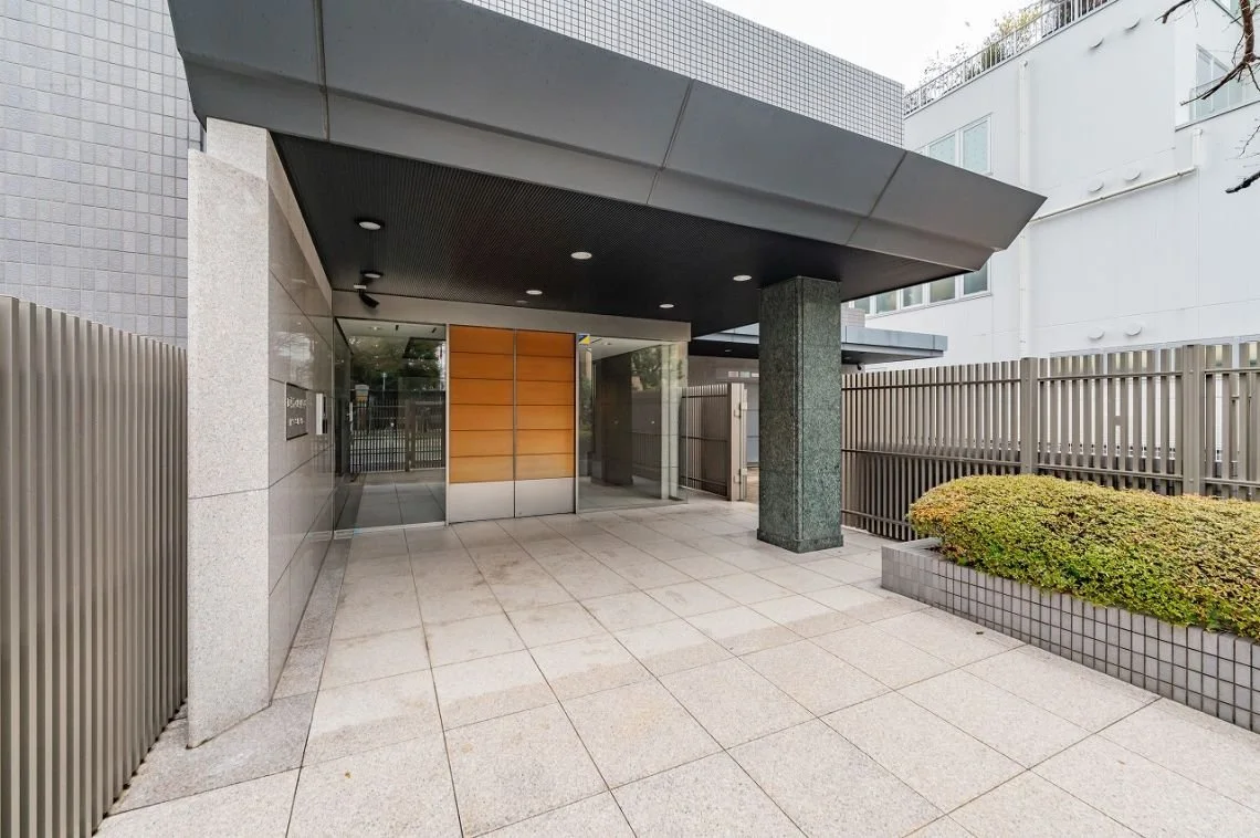 モダンな建物の入り口、ガラスの扉と木製のドア、コンクリートの柱とフェンスがあります。