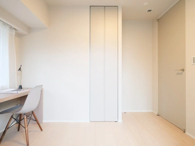 白い壁の部屋に、左側に机と椅子、中央に縦長の収納棚、右側にドアと窓。