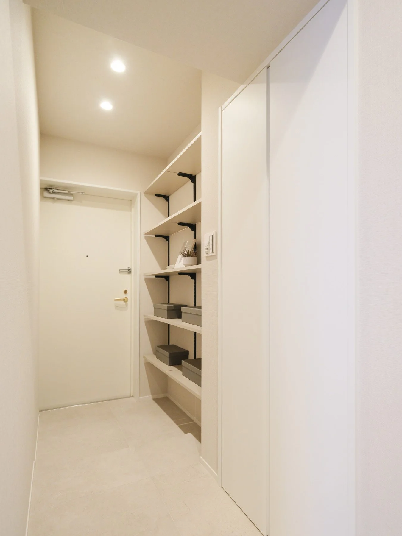 白い壁と扉、棚に灰色の箱や装飾品が置かれたシンプルな玄関・廊下の interior.