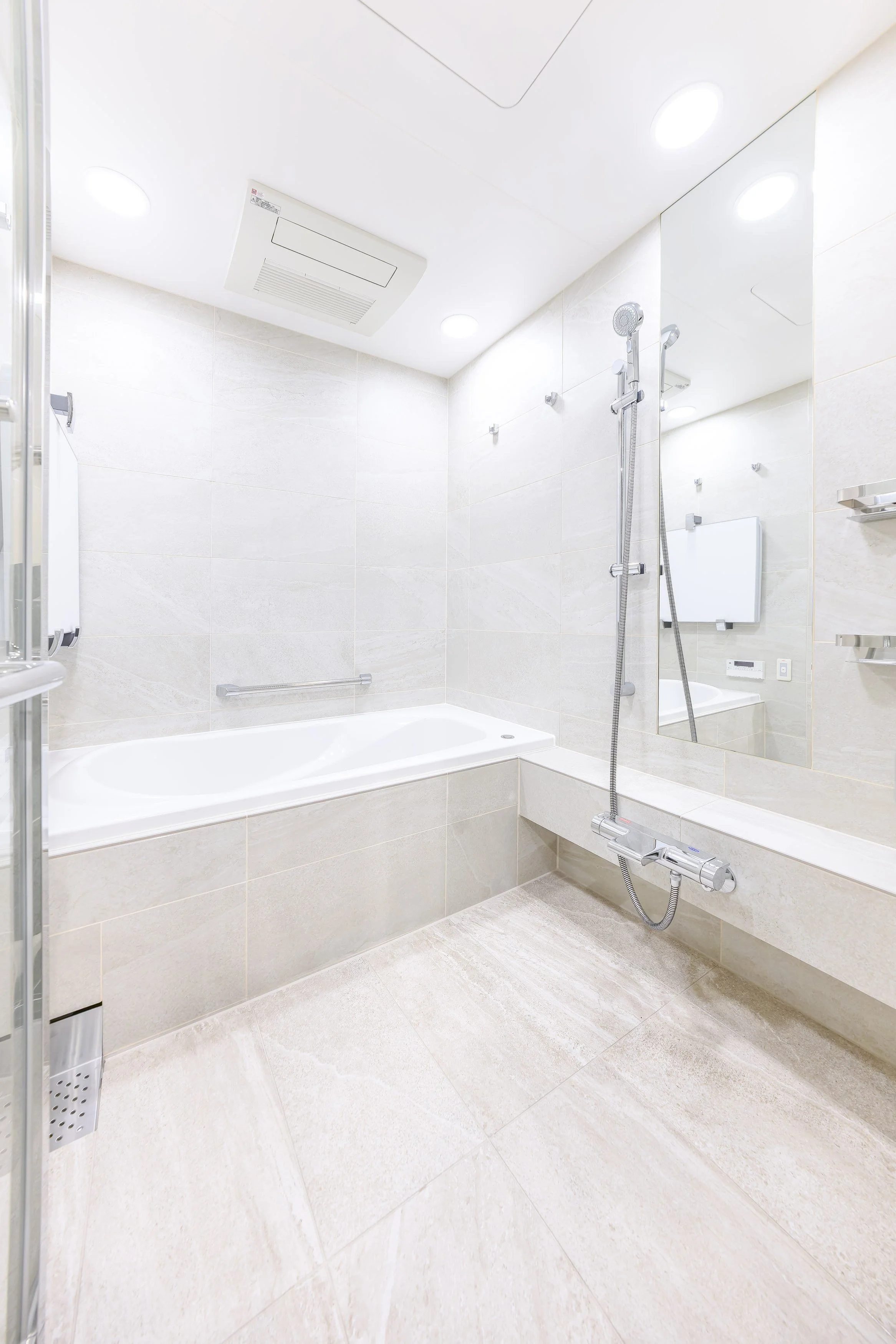 白い浴槽とシャワーヘッドがついたバスルーム。壁に鏡と棚があり、天井には照明と換気扇が設置されている。