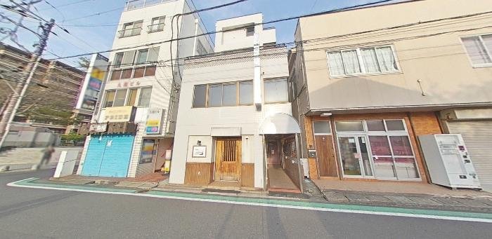 三階建ての白い建物と隣接する建物の街並み。道路沿いに並ぶ店や建物の外観。