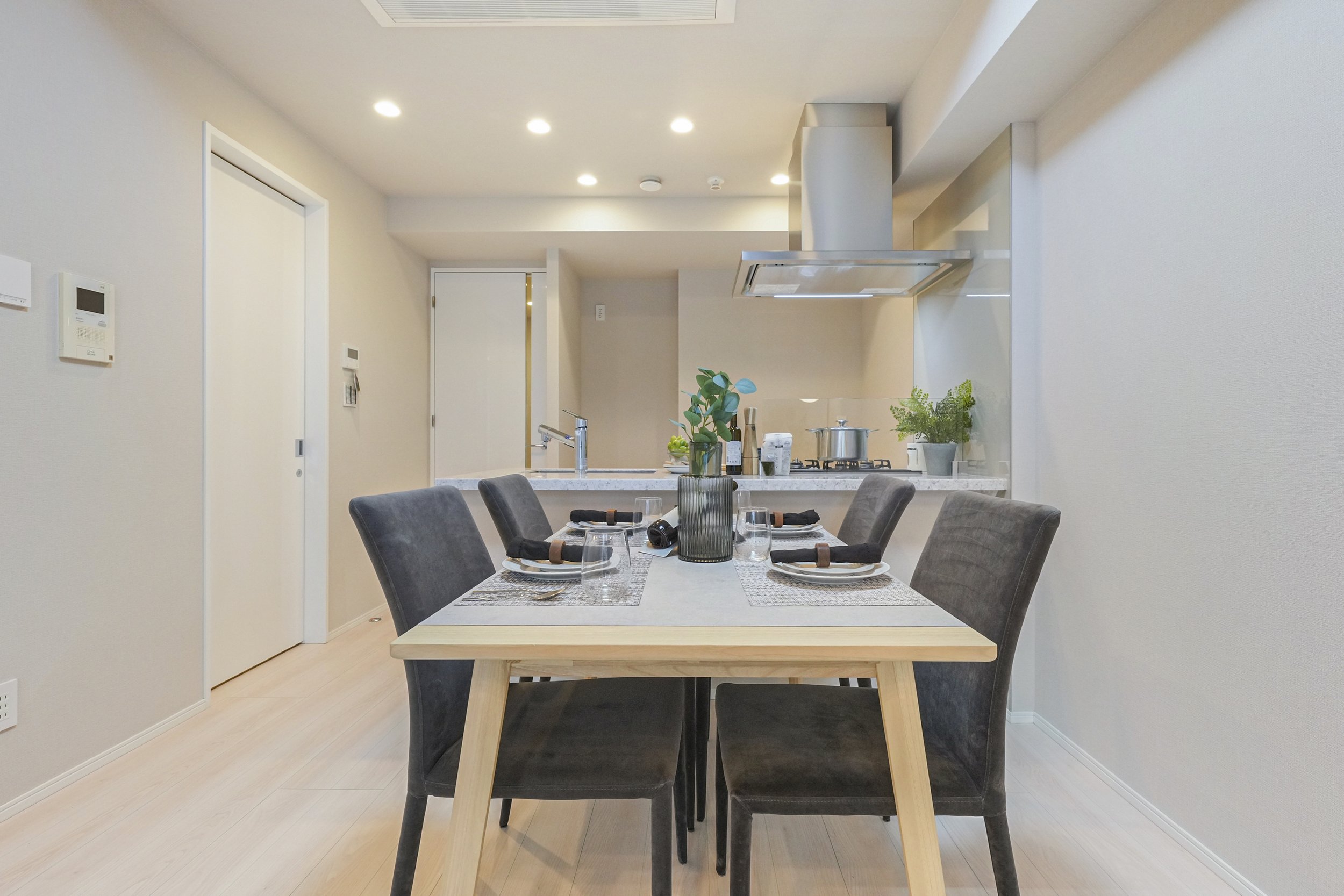 ダイニングテーブルにセットされた食器とグラス、花瓶に入った植物、キッチンエリアのガスコンロと調理器具、室内の明るいリビングルームの画像