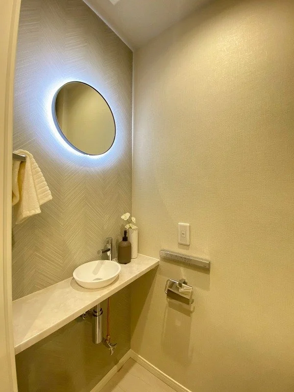 シンプルなトイレの洗面台と丸い鏡、壁には暖色の照明が付いている