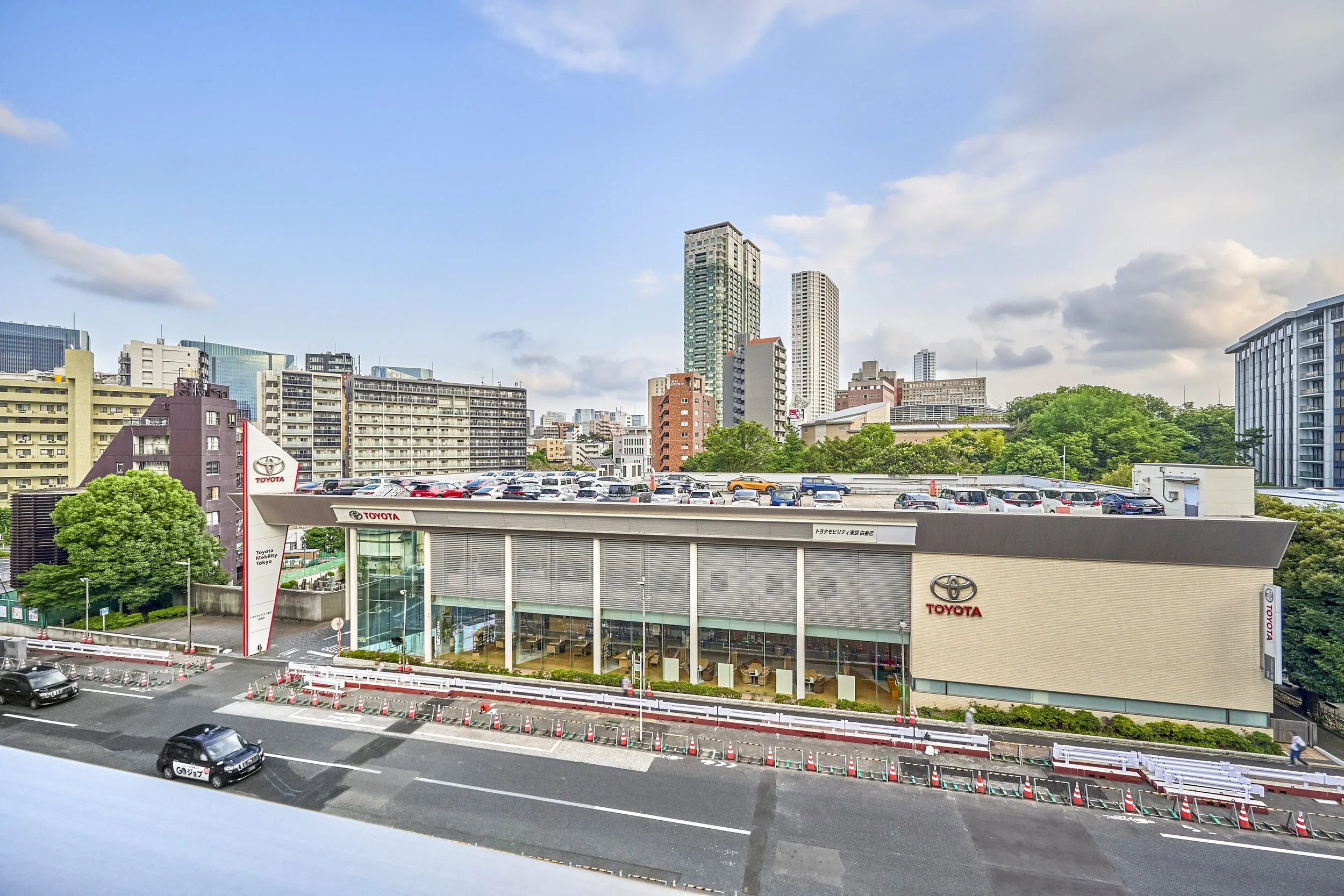 東京の都市景観、ビルと高層ビルが立ち並び、駐車場と道路も見える。青空に白い雲が浮かぶ。