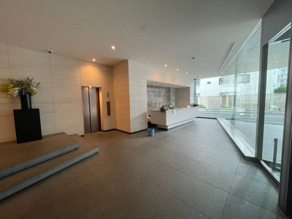 ホテルのロビーの入り口付近の景色。エレベーターと受付カウンターがあり、花の飾りが置かれている。大きなガラス窓から外の景色が見える。