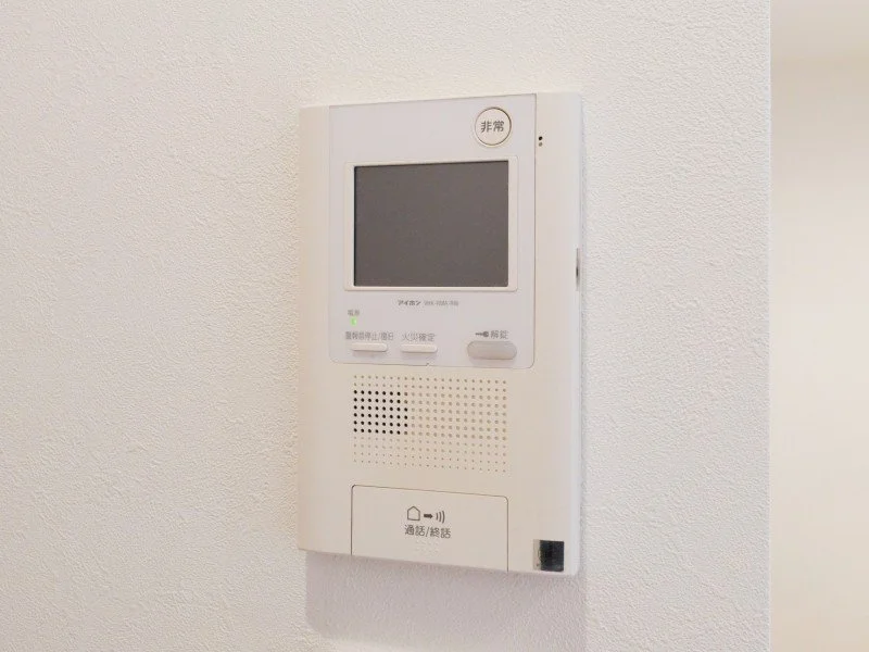 壁に取り付けられた白いインターホンまたはセキュリティモニターの装置。小さな画面とボタンが見える。