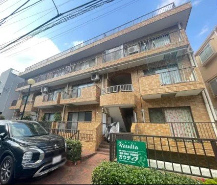 四階建てのアパートメントの建物と駐車場。駐車中の車と緑の看板が見える。