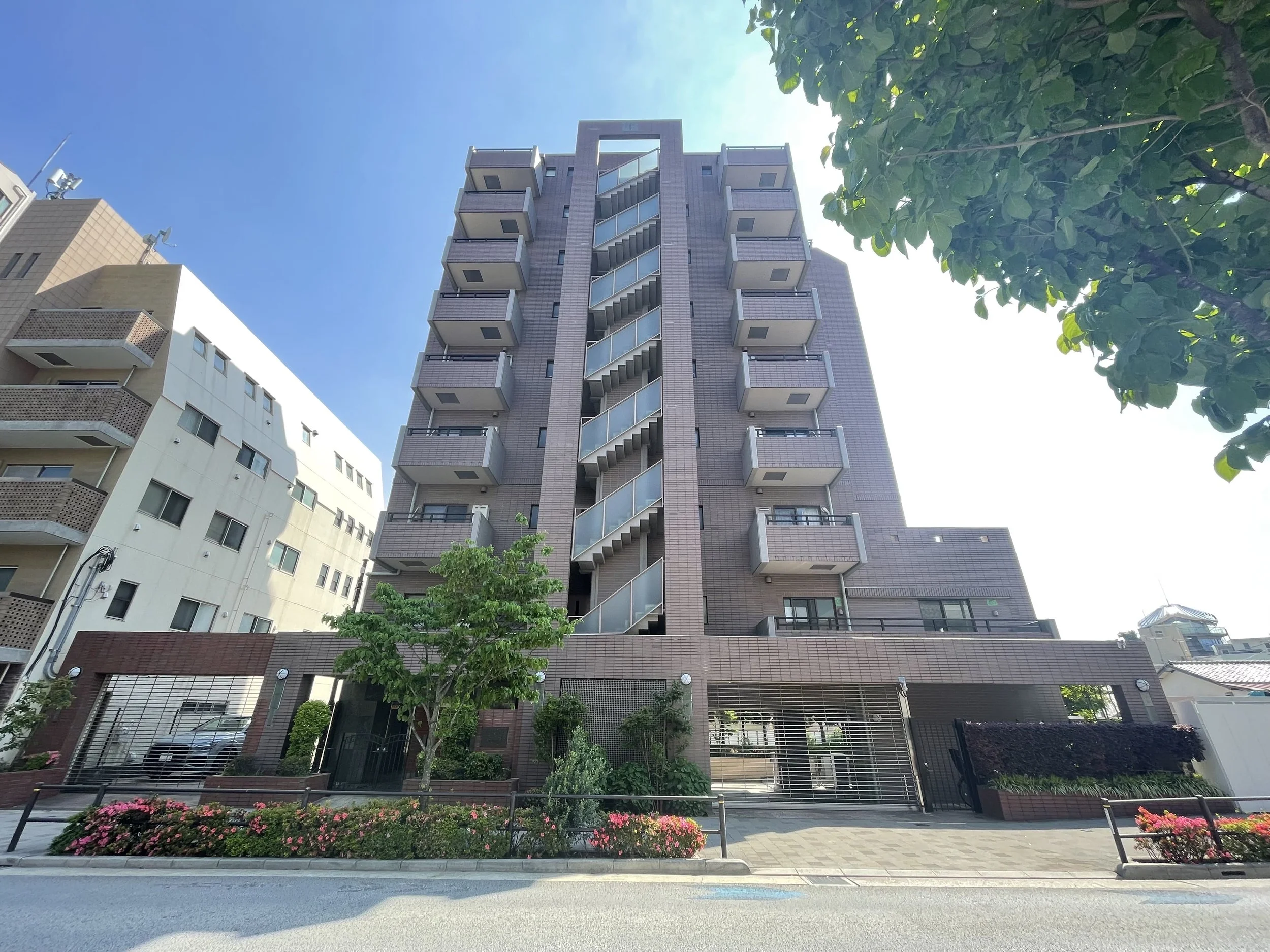 中層のマンションと周囲の建物、庭の花と木々、青空と一部の葉