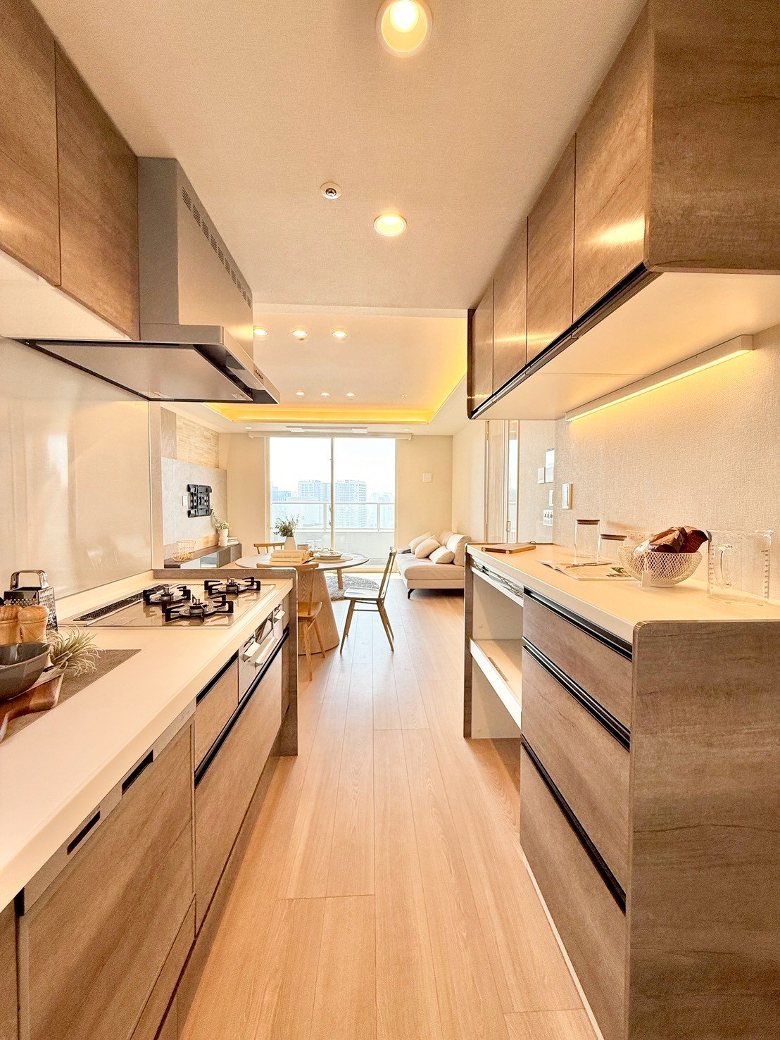 モダンなキッチンとリビングルーム。木製の床とキャビネット、天井には埋め込み照明。後ろにはソファと大きな窓からの景色。