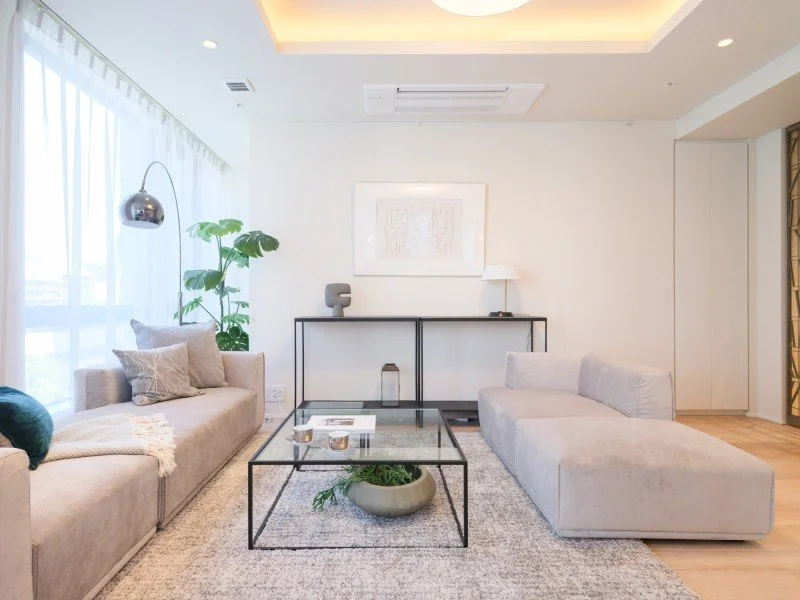 リビングルームのインテリア、白いソファ、ガラスのコーヒーテーブル、観葉植物、壁にアートワーク、自然光が差し込む窓