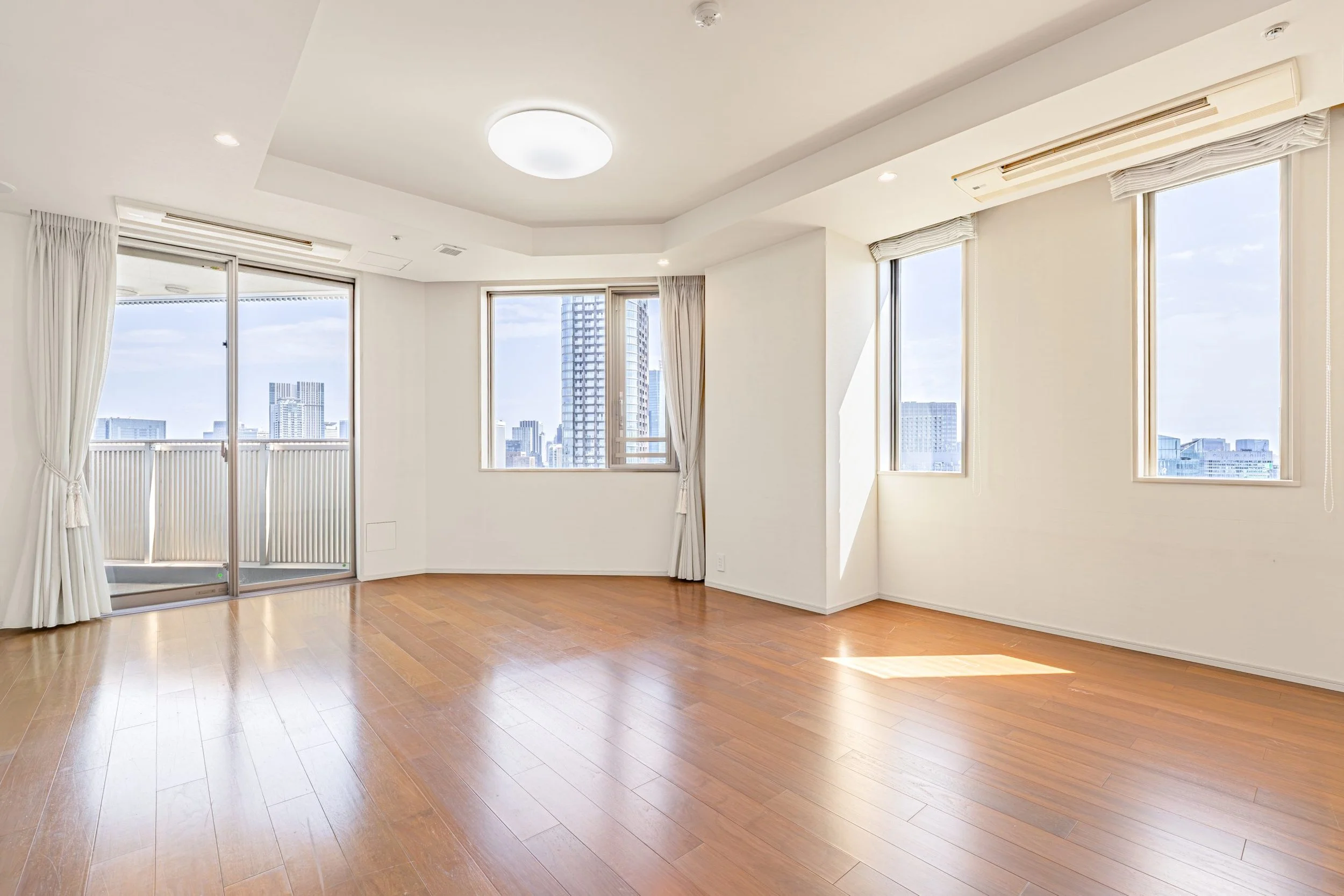 白い壁と木製フローリングのリビングルーム、窓から都市の高層ビル群が見える