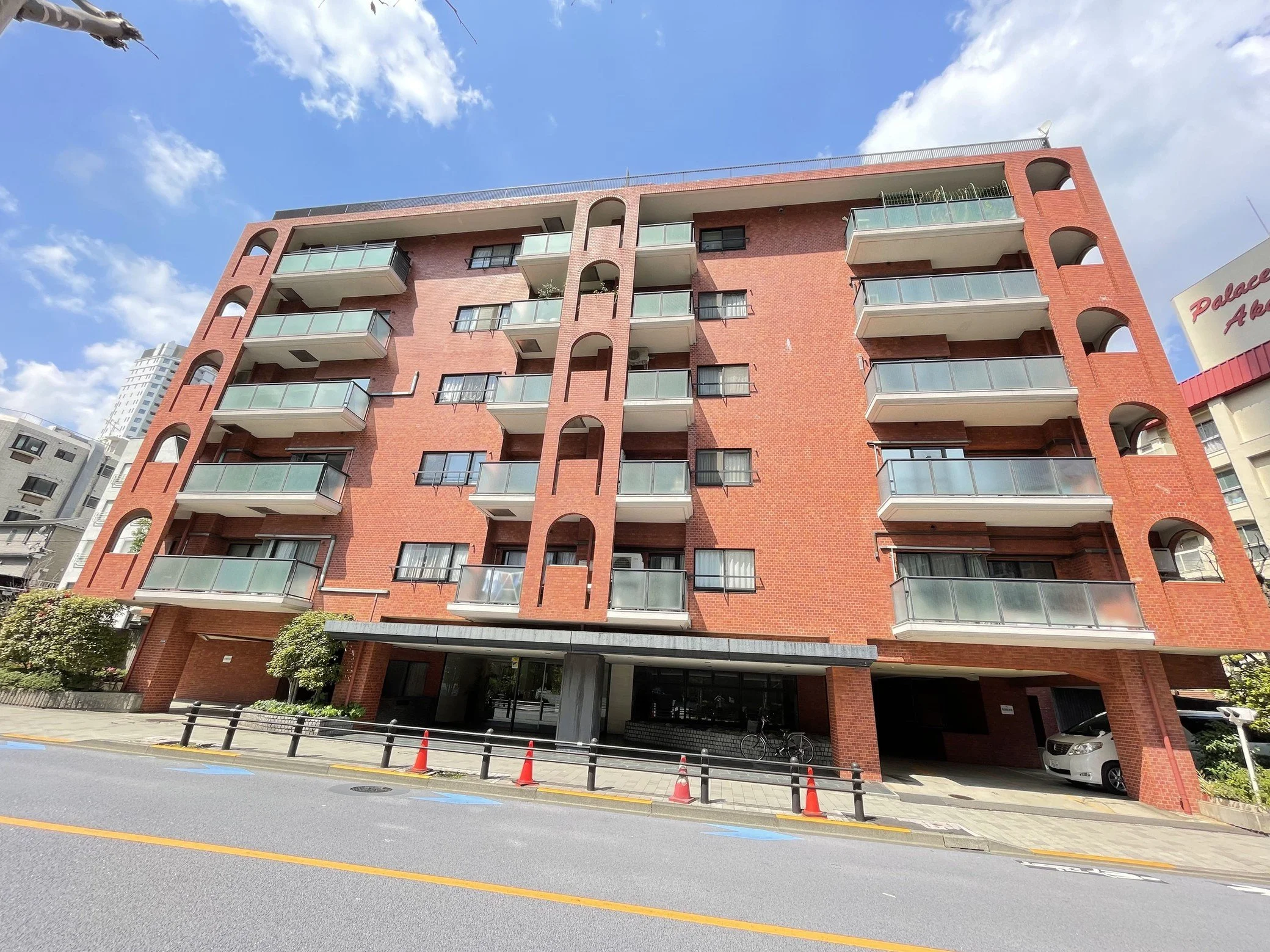 赤いレンガ造りの中層マンション。前面道路と歩道が見える。空は青く雲が散らばる晴天。