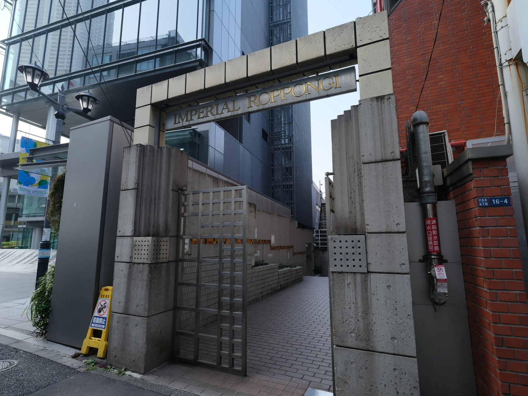 東京のビル街にある入り口の門で、上部に「IMPERIAL ROPPONGI」と書かれた金色の文字がある。石とコンクリートの構造物と金属製の門とチェーンが設置されている。周囲に街灯や看板、建物のガラス面が見える。