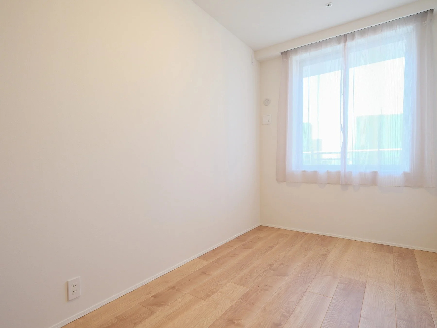 白い壁と窓がある空き部屋。木製の床とカーテンが掛かった窓から自然光が入っている。