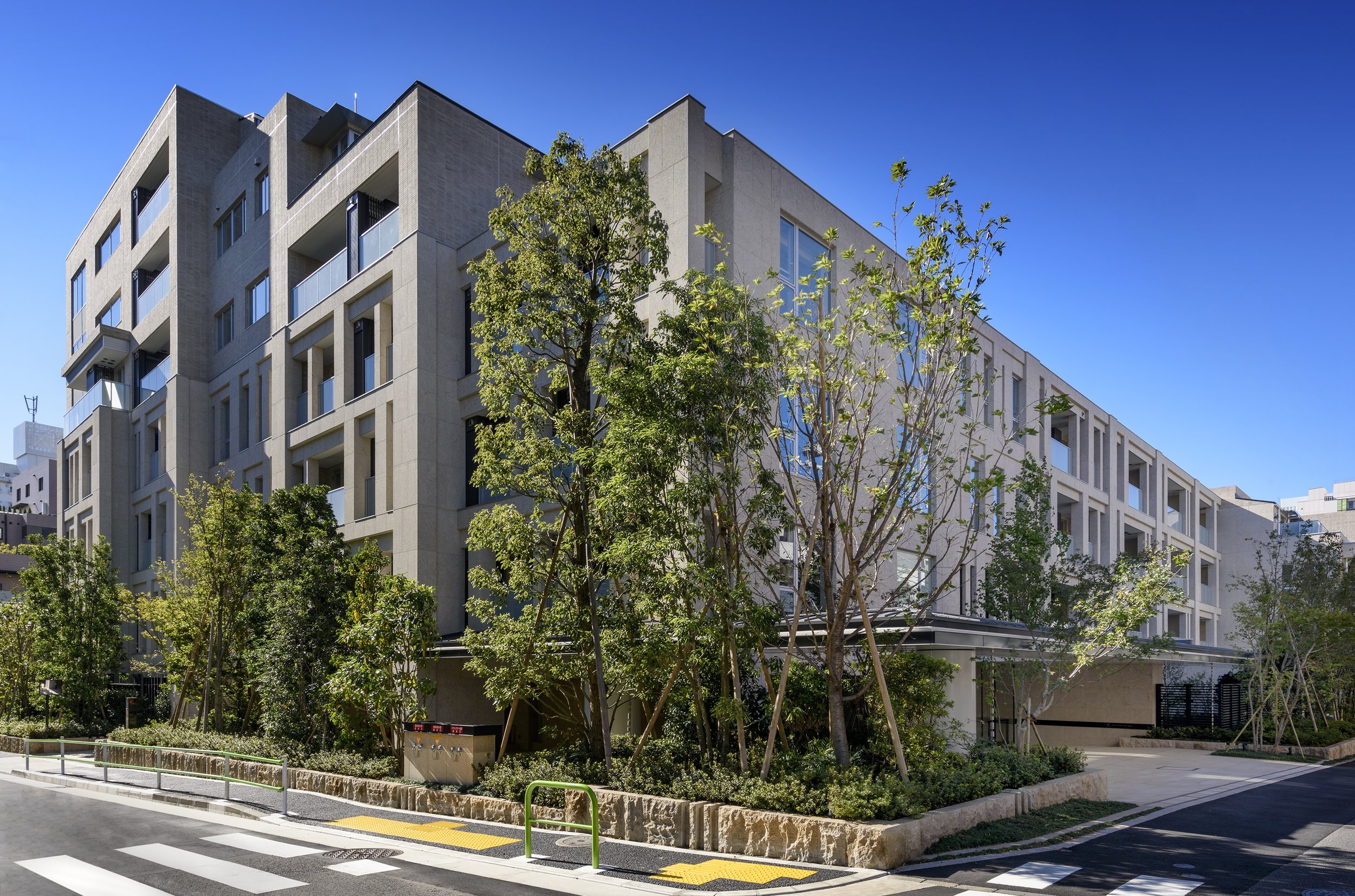 高層のコンクリート造りの住宅またはアパートメントの建物、前景に木と低木の植え込み、道路と横断歩道がある。青空が背景。