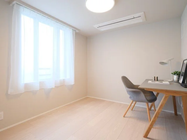 白い壁とカーテンを通して陽光が入るシンプルな書斎部屋。机の上に書類と植物が置かれ、椅子に座る場所。