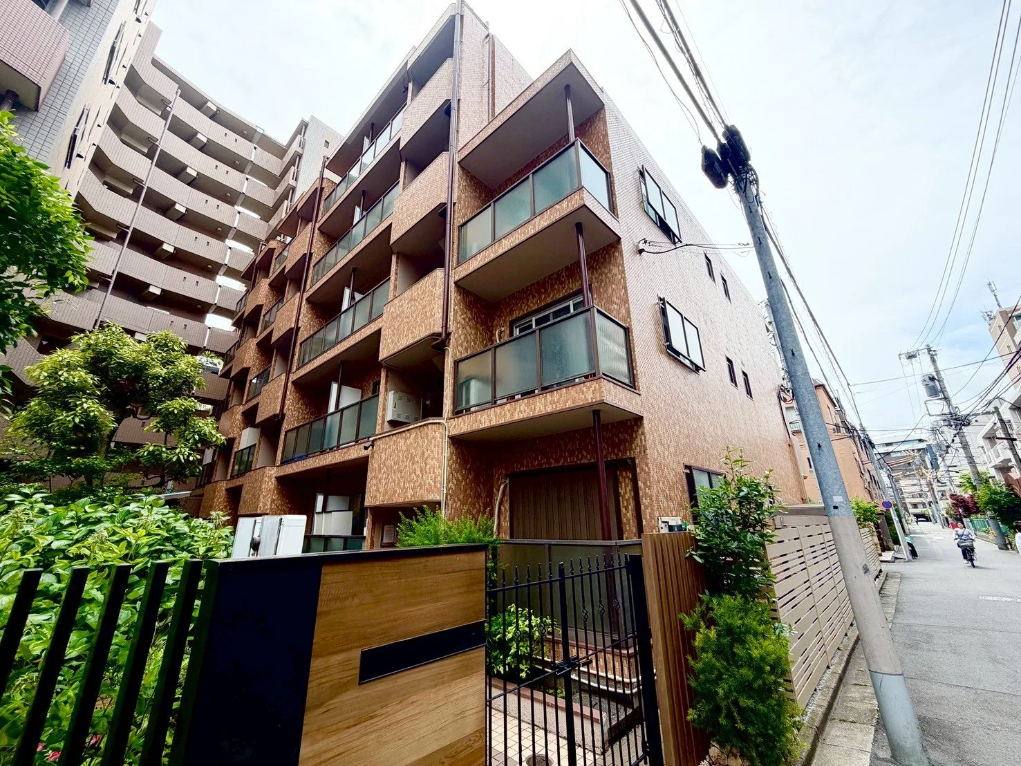 多層階の茶色いマンションと道路に電柱。前に緑の植物とフェンスがある。