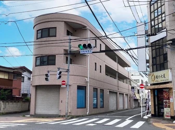 三階建ての角にあるピンクのレンガ造りの建物。道路の交差点にあり、セブンイレブンと飲食店の看板も見える。上空には電線が交差している。