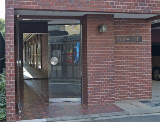 赤レンガの建物の入り口。左側はガラス扉で、中には通路と店舗の入口が見える。右側に看板があり、外壁に照明が設置されている。