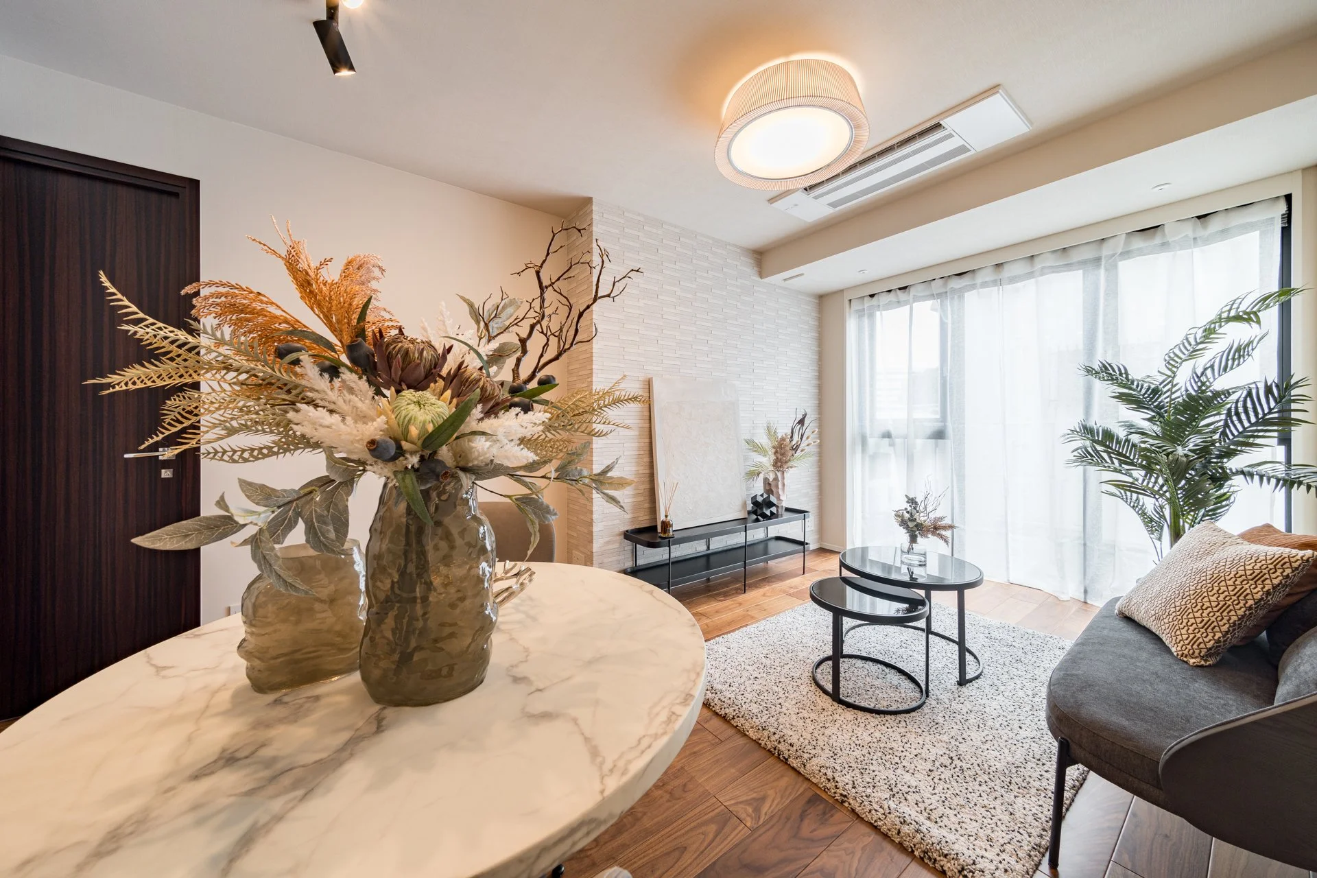 リビングルームのインテリア。丸い大理石のテーブルに花瓶と花の飾り、背景にソファとコーヒーテーブル、窓から自然光が差し込み、観葉植物も配置されている。