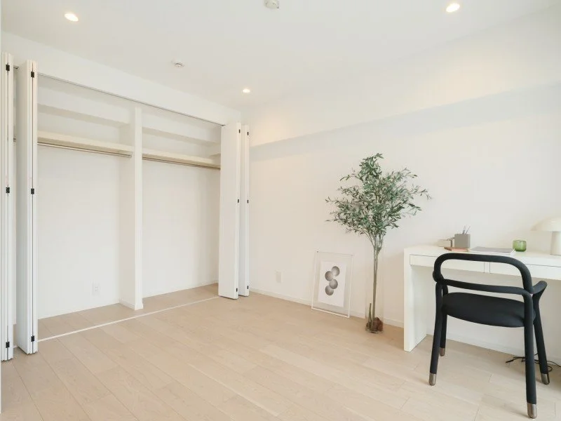 白い壁の部屋に、空のクローゼットと机と椅子、観葉植物が置かれているシンプルな室内風景