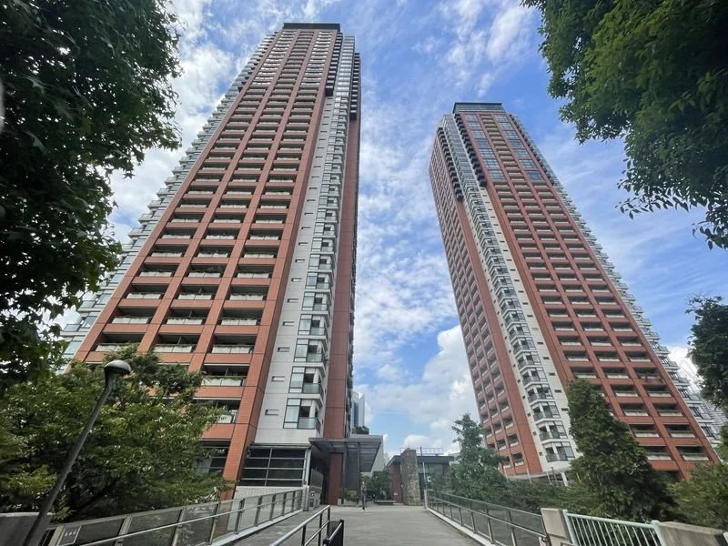 青空を背景に並んで建つ赤茶色の高層マンション2棟と、木々に囲まれた歩道