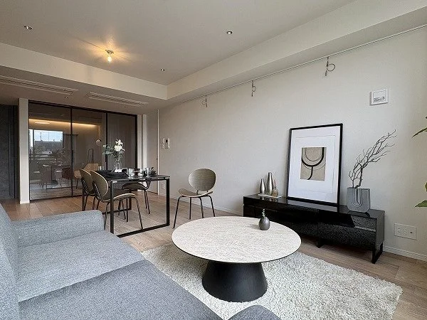 モダンなリビングルームの室内写真、ソファ、円形のコーヒーテーブル、壁にアート、ダイニングテーブルと椅子、バルコニーのガラス扉が見える