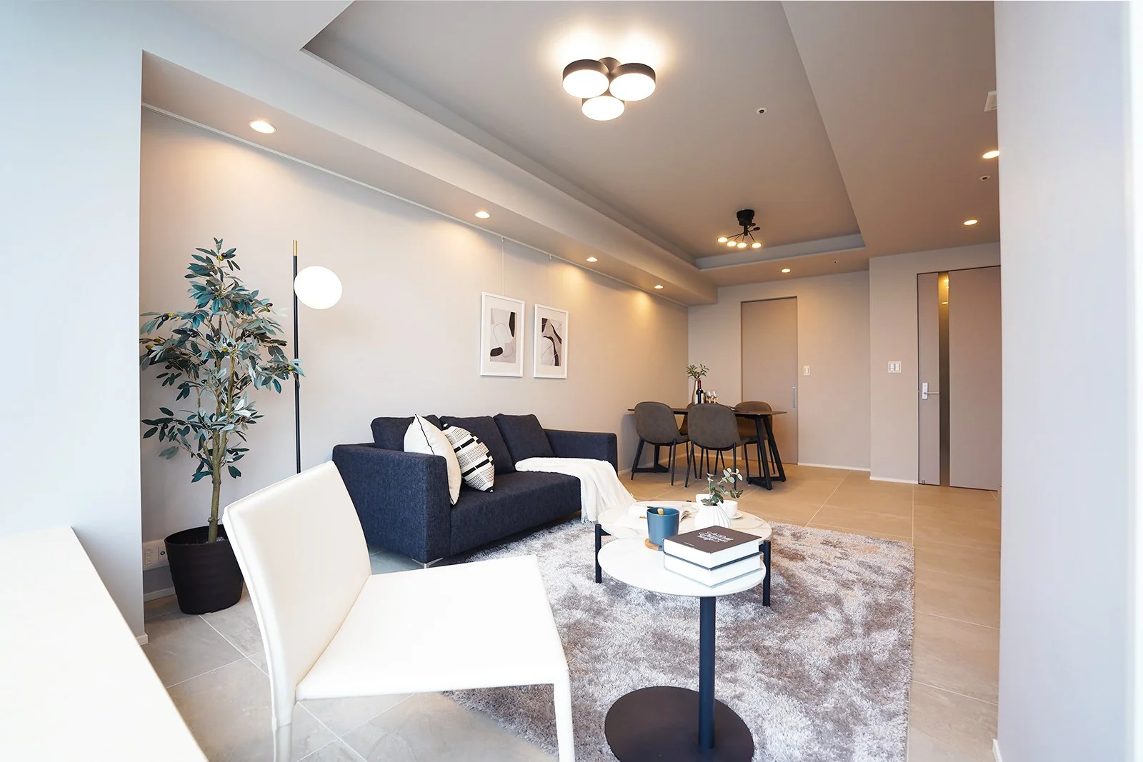 シンプルでモダンなリビングルーム、黒いソファ、白い椅子、丸いコーヒーテーブル、観葉植物、壁に2つの抽象画、ダイニングテーブルと椅子、間接照明と天井のライト