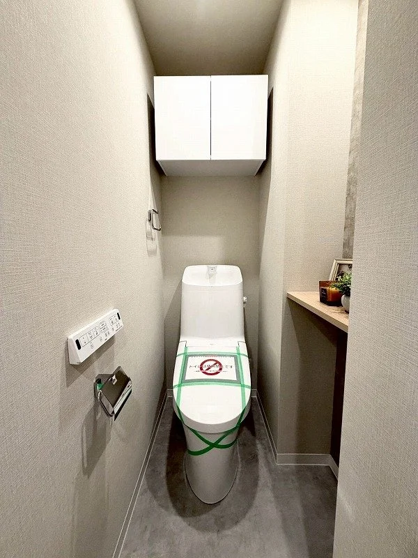 トイレの便器と壁に取り付けられた収納棚、壁に設置されたコントロールパネルとトイレットペーパーホルダー、横に棚と飾り棚がある小さなトイレルーム